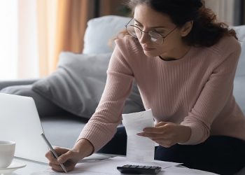 Focused millennial woman in eyewear writes out information from invoices, calculating monthly expenses alone at home. Young lady managing incomes and outcomes, planning investments or summer vacation.