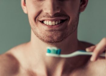 Cropped image of handsome young man with bare torso holding a toothbrush and smiling, on gray background