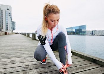Young woman tying her shoelaces before a run along waterfront. Female runner preparing foe sprint. Fit female athlete on boardwalk along river.