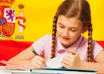 Diligent teenage student, Caucasian girl, writing with a pen in her copybook, Spanish flag behind